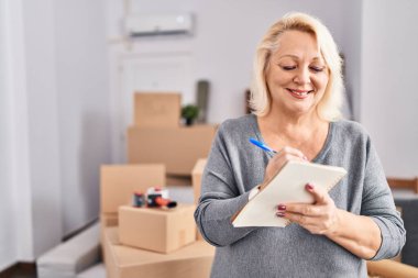 Middle age blonde woman smiling confident writing on notebook at new home