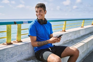 Genç Latin adam sahilde kulaklık ve akıllı telefon kullanarak mutlu gülümsüyor..