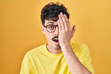 Hispanic man wearing glasses standing over yellow background covering one eye with hand, confident smile on face and surprise emotion. 