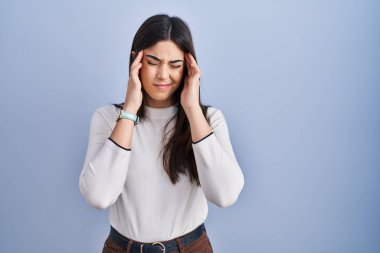 Mavi arka planda duran genç esmer kadın, elleri başının üstünde, stresten baş ağrısı çekiyor. migren ağrısı. 