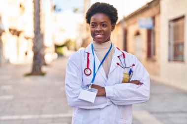Doktor üniforması giyen Afrikalı Amerikalı kadın caddede kollarını kavuşturmuş duruyor.