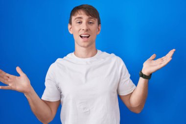 Caucasian blond man standing over blue background celebrating crazy and amazed for success with arms raised and open eyes screaming excited. winner concept 