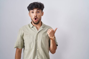 Arab man with beard standing over white background surprised pointing with hand finger to the side, open mouth amazed expression. 