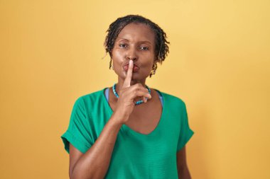 African woman with dreadlocks standing over yellow background asking to be quiet with finger on lips. silence and secret concept. 