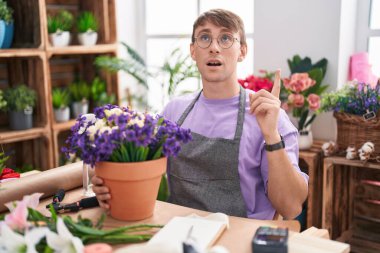 Caucasian blond man working at florist shop amazed and surprised looking up and pointing with fingers and raised arms. 