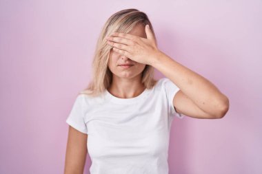 Young blonde woman standing over pink background covering eyes with hand, looking serious and sad. sightless, hiding and rejection concept 