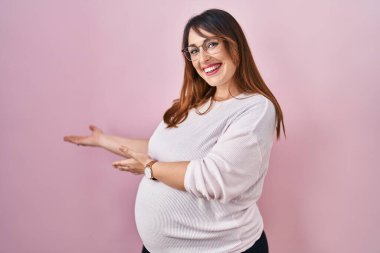 Pregnant woman standing over pink background inviting to enter smiling natural with open hand 