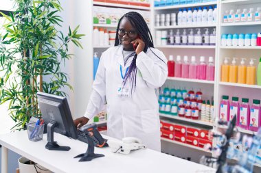 Afrika kökenli Amerikalı kadın eczacı eczanedeki bilgisayarla akıllı telefondan konuşuyor.