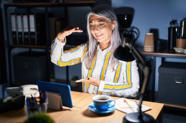 Gri saçlı, geceleri ofiste çalışan orta yaşlı bir kadın. Ellerinde büyük ve büyük boy işareti, ölçü sembolü olan bir el hareketi yapıyor. Kameraya bakıp gülümsüyor. ölçüm kavramı. 