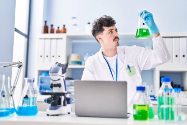 Young caucasian man scientist using laptop holding test tube at laboratory