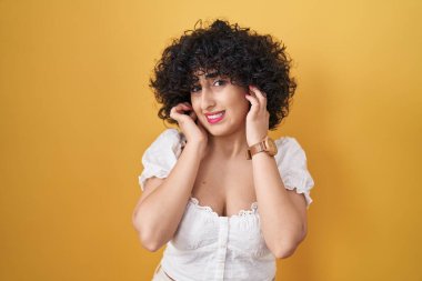 Sarı arka planda duran kıvırcık saçlı genç esmer kadın yüksek sesli müzik sesi için kulaklarını parmaklarıyla kapatıyor. sağır kavramı. 