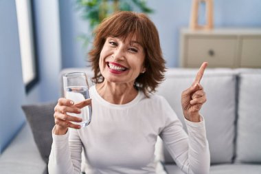 Orta yaşlı kadın bir bardak su içiyor. Gülümsüyor, mutlu bir şekilde elini ve parmağını yana doğru gösteriyor. 