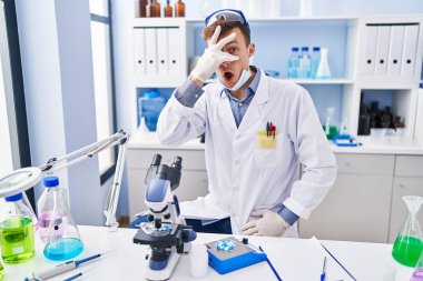 Caucasian man working at scientist laboratory peeking in shock covering face and eyes with hand, looking through fingers afraid 