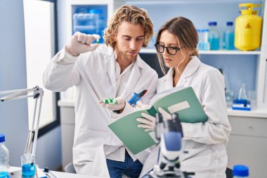 Man and woman wearing scientist uniform holding pills writing on notebook at laboratory