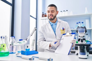 Hispanic man working at scientist laboratory holding blank clipboard winking looking at the camera with sexy expression, cheerful and happy face. 
