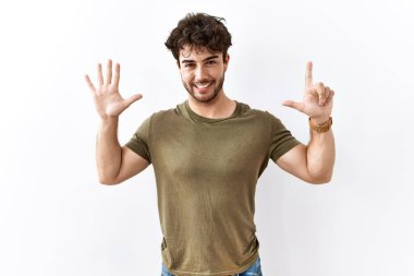 İzole edilmiş beyaz arka planda duran İspanyol adam yedinci parmaklarını gösterip işaret ederken kendinden emin ve mutlu gülümsüyor.. 