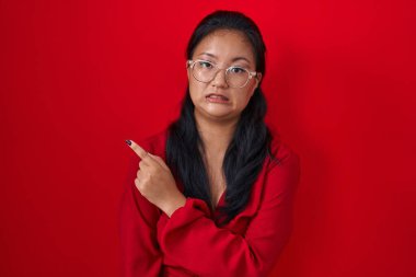 Asian young woman standing over red background pointing aside worried and nervous with forefinger, concerned and surprised expression 