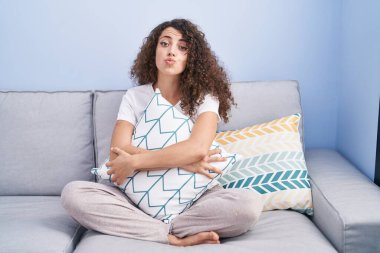 Hispanic woman with curly hair sitting on the sofa at home looking at the camera blowing a kiss being lovely and sexy. love expression. 