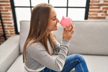 Young blonde woman smiling confident holding piggy bank at home
