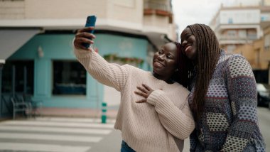 İki Afrikalı Amerikalı arkadaş gülümsüyor. Kendilerine güveniyorlar. Sokakta akıllı telefonun yanında selfie çekiyorlar.