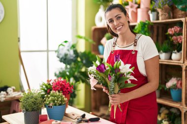 Young beautiful hispanic woman florist holding bouquet of flowers at flower shop