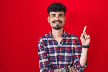 Kırmızı arka planda duran sakallı, yüzünde kocaman bir gülümseme olan, eli kameraya bakan genç İspanyol bir adam.. 