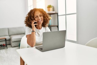 Orta yaşlı İspanyol kadın evde akıllı telefondan dizüstü bilgisayar kullanıyor.