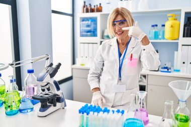 Middle age blonde woman working at scientist laboratory pointing with hand finger to face and nose, smiling cheerful. beauty concept 