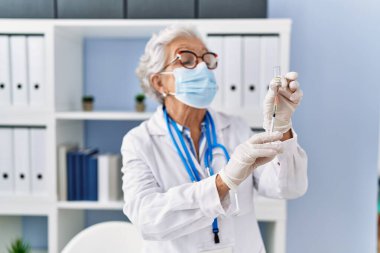 Doktor üniforması giyen yaşlı gri saçlı kadın ve klinikte aşıyı elinde tutan tıbbi maske.