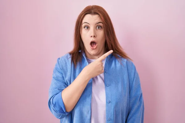 Young hispanic woman with red hair standing over pink background surprised pointing with finger to the side, open mouth amazed expression.