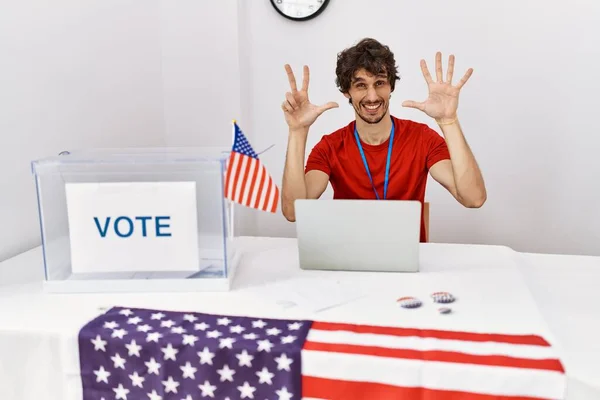 Young hispanic man at political election sitting by ballot showing and pointing up with fingers number eight while smiling confident and happy.