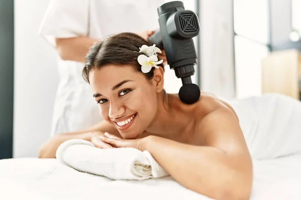 Young latin woman relaxed having back massage with percussion pistol at beauty center