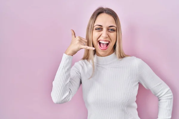 Young blonde woman wearing white sweater over pink background smiling doing phone gesture with hand and fingers like talking on the telephone. communicating concepts.