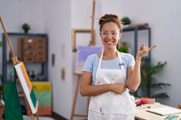 Brunette woman painting at art studio smiling happy pointing with hand and finger to the side