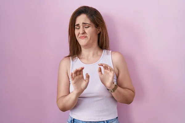 Brunette woman standing over pink background disgusted expression, displeased and fearful doing disgust face because aversion reaction.