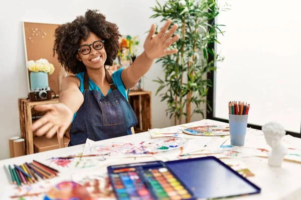 Beautiful african american woman with afro hair painting at art studio looking at the camera smiling with open arms for hug. cheerful expression embracing happiness.