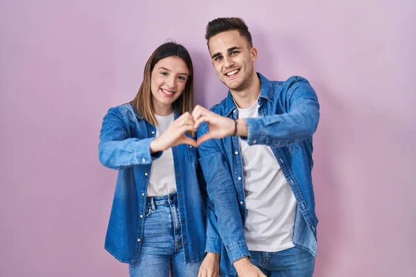 Young Hispanic Couple Standing Pink Background Smiling Love Doing Heart — Stockfoto