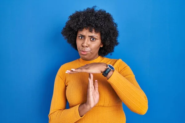 Black woman with curly hair standing over blue background doing time out gesture with hands, frustrated and serious face