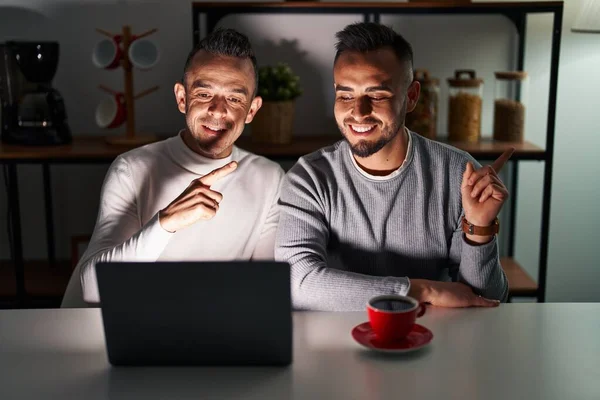 Homosexual couple using computer laptop with a big smile on face, pointing with hand finger to the side looking at the camera.