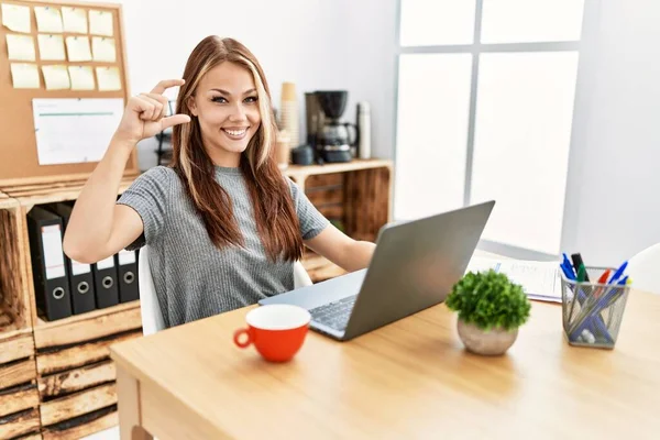 在办公室工作的年轻的黑发女人 带着笔记本电脑微笑 自信地摆姿势 手拿着小尺寸的标志 手指看着相机 计量概念 — 图库照片