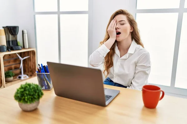 Young caucasian woman working at the office using computer laptop yawning tired covering half face, eye and mouth with hand. face hurts in pain.