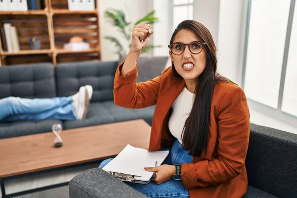 Young hispanic woman working as psychology counselor angry and mad raising fist frustrated and furious while shouting with anger. rage and aggressive concept.