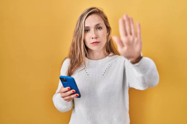 スマートフォンの入力メッセージを使用して若いブロンドの女性が手の手のひらで歌うのを停止します 顔に負の重大なジェスチャーで — ストック写真