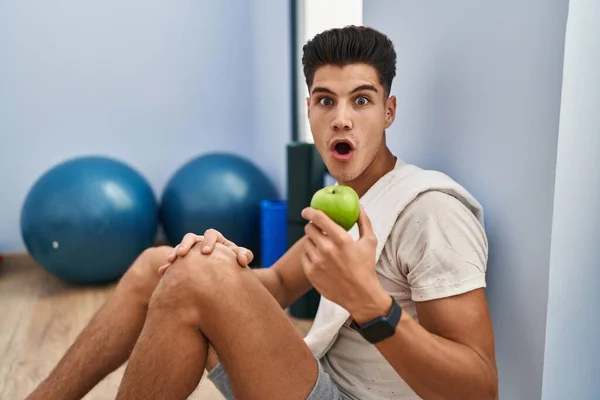 若いヒスパニック系の男は健康的なリンゴを食べスポーツウェアを身に着けている恐怖と驚きのために口を開けて驚いて 不信の顔 — ストック写真