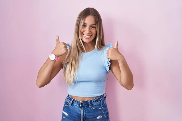 Young blonde woman standing over pink background success sign doing positive gesture with hand, thumbs up smiling and happy. cheerful expression and winner gesture.