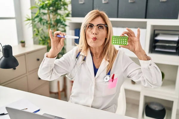 Middle age blonde doctor woman holding prescription pills and pregnancy test making fish face with mouth and squinting eyes, crazy and comical.