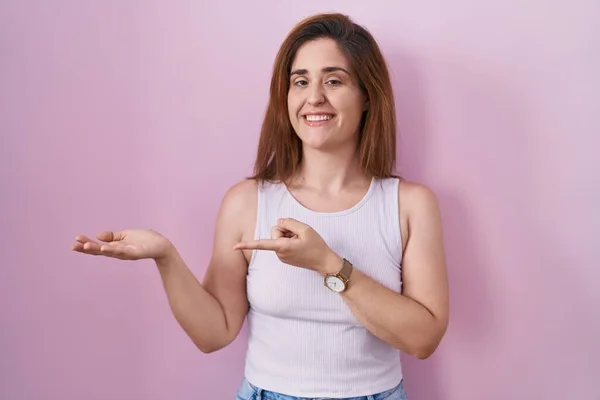 Brunette Woman Standing Pink Background Amazed Smiling Camera While Presenting — Zdjęcie stockowe