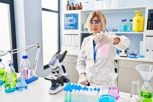 Middle age blonde woman working at scientist laboratory looking unhappy and angry showing rejection and negative with thumbs down gesture. bad expression.