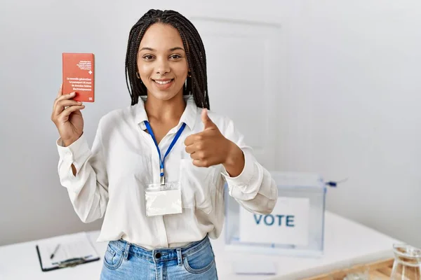 Young african american woman at political campaign election holding swiss passport smiling happy and positive, thumb up doing excellent and approval sign