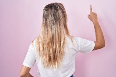 Young blonde woman standing over pink background posing backwards pointing ahead with finger hand 